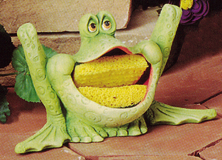 A cast ceramic figure of a seated, cartoonishly styled frog with a wide open mouth. The mouth opens to show the inside of the figure is hollow, and is sized to hold a kitchen sponge inside it.