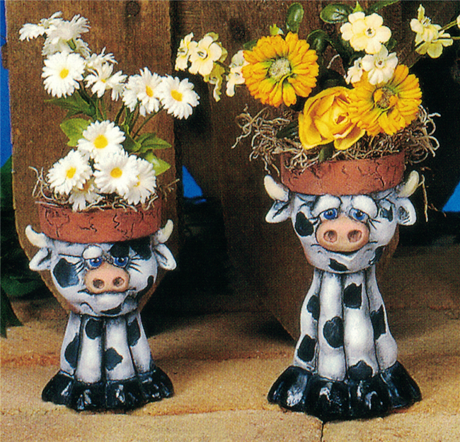 Two separate small cast ceramic figurines of cartoonish cows. Their heads have each been hollowed out and topped with a rim like a planter pot. The left cow is shorter and stockier, the right taller and thinner. Each stands atop four legs with wide hooves