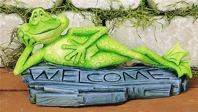 A cast ceramic figure of a cartoonishly styled frog laying on its side atop a worn plank of wood. The frog supports its head with one arm and has its ankles crossed. The word WELCOME is carved into the plank below it as it looks forward with a smile.
