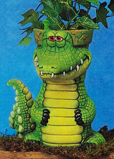 A cast ceramic pot that looks like a cartoonish alligator. The back of its head is the pot, with the rim just visible behind its eyes. It is seated with its tail curved to point up by its side. Its hands wrap around its belly.