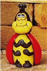 A ceramic figure of a cartoonishly styled ladybug. It is seated with its front two pairs of arms held to its chest. It looks upwards smiling, its wings around its shoulders like a cape. Its antennae are short and stocky.