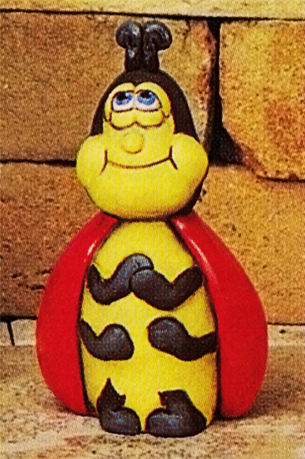 A ceramic figure of a cartoonishly styled ladybug. It is seated with its front two pairs of arms held to its chest. It looks upwards smiling, its wings around its shoulders like a cape. Its antennae are short and stocky.