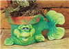 A small cast ceramic pot made to resemble a cartoonish fish. Its head and body form the pot, with the rim visible above its eyes. It lies on its stomach, fins extended, looking forward with a small grin. Its tail extends out to the side.