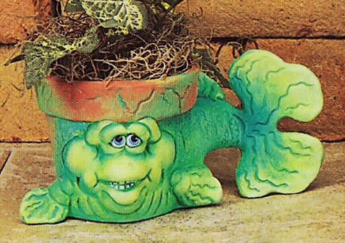 A small cast ceramic pot made to resemble a cartoonish fish. Its head and body form the pot, with the rim visible above its eyes. It lies on its stomach, fins extended, looking forward with a small grin. Its tail extends out to the side.