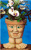 A cast ceramic pot with a cartoonish human face. The face has an open mouth smile, large nose, lidded eyes, and round cheeks. The pot is textured with small lines like aged cracks and is placed atop a pair of wide cartoonish cast ceramic feet.