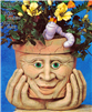 A cast ceramic pot with a cartoonish human face. The face has an open mouth smile, large nose, and lidded eyes. The pot is textured with small lines like aged cracks and is placed atop a pair of cartoonish cast ceramic hands as if it was holding its chin.
