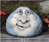 A cast ceramic figure of a rock with a cartoonish human face. It is shaped somewhat like a gumdrop. It has two wrinkled eyes, a very large nose, round cheeks, and a smile with lips slightly open and to the left.