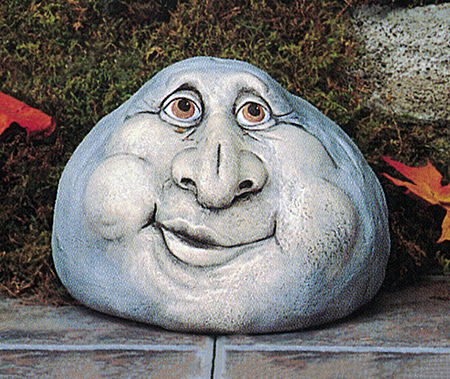 A cast ceramic figure of a rock with a cartoonish human face. It is shaped somewhat like a gumdrop. It has two wrinkled eyes, a very large nose, round cheeks, and a smile with lips slightly open and to the left.