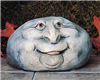 A cast ceramic figure of a rock with a cartoonish human face. It is somewhat oval in shape. It has two wide set wrinkled eyes, a large nose, round cheeks and chin, and a smile with a slightly visible tongue poking out.