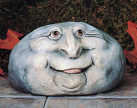 A cast ceramic figure of a rock with a cartoonish human face. It is somewhat oval in shape. It has two wide set wrinkled eyes, a large nose, round cheeks and chin, and a smile with a slightly visible tongue poking out.