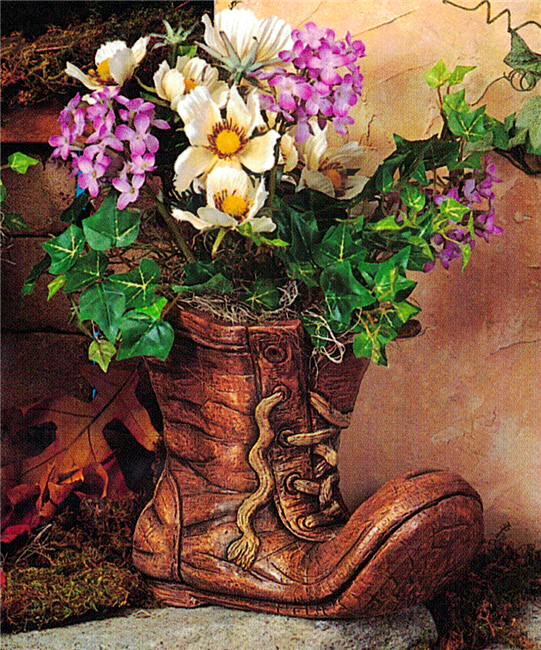 A cast ceramic planter shaped like a worn boot. The toe of the boot is bent upwards and its laces are undone and frayed at the ends. It is wrinkled and painted to resemble old leather. It is filled with real flowers and greenery.
