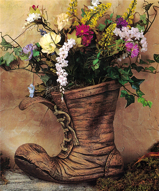 A large cast ceramic planter shaped like a worn boot. The toe of the boot is bent upwards and its laces are undone and frayed at the ends. It is wrinkled and painted to resemble old leather. It is filled with real flowers and greenery.