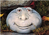 A cast ceramic figure of a rock with a cartoonish human face. It is somewhat oval in shape. It has two round eyes, a large nose, round cheeks, and wears a calm, placid smile.