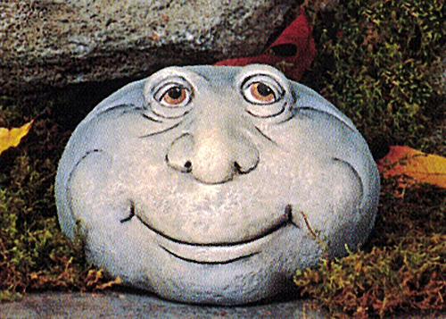 A cast ceramic figure of a rock with a cartoonish human face. It is somewhat oval in shape. It has two round eyes, a large nose, round cheeks, and wears a calm, placid smile.