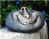 A cast ceramic figure of a rock with a cartoonish human face. It is somewhat oval in shape. It has two deep set eyes and a wide, mischievous grin with a few visible teeth.