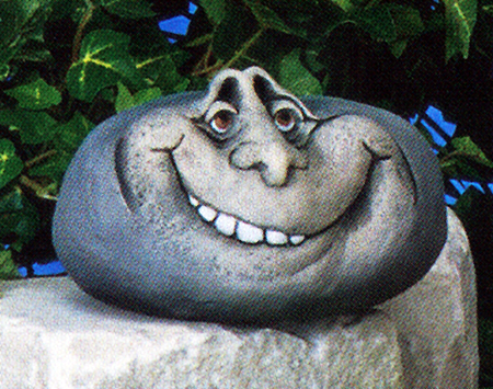 A cast ceramic figure of a rock with a cartoonish human face. It is somewhat oval in shape. It has two deep set eyes and a wide, mischievous grin with a few visible teeth.