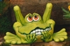 A cast ceramic statue of a cartoonish frog. It sits, bent back legs protruding well above its head. It looks up with large eyes and a nervous, toothy mouth. Its hands are to its mouth as if chewing its nails.