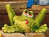 A cast ceramic statue of a cartoonish frog. It sits, bent back legs protruding well above its head. It looks upward with lidded eyes, its head tilted. It sticks its tongue out slightly, and holds its face in its hands.