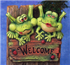 A cast ceramic plaque featuring two cartoonish frogs seated atop a wooden sign reading Welcome. The left frog has its tongue out slightly and looks to the side. The left wears lipstick and looks forward. There are three ladybugs also on the sign.