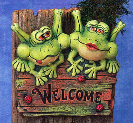 A cast ceramic plaque featuring two cartoonish frogs seated atop a wooden sign reading Welcome. The left frog has its tongue out slightly and looks to the side. The left wears lipstick and looks forward. There are three ladybugs also on the sign.