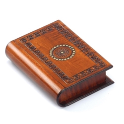 This trick box is shaped like a book and decorated with a spiral border pattern around a floral circle accented with gold paint. The binding of the book slides to reveal a removable panel allowing access to the box compartment.