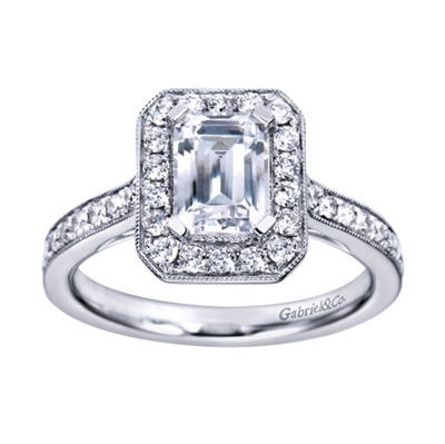 An emerald cut center diamond gleams magnificently encircled in round brilliant diamonds set in a vintage halo engagement ring designed by Gabriel & Co.