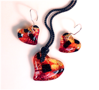 Three heart-shaped pendants made from fused glass. Each is made from pieces of red, orange, and yellow glass with a few chunks of black. The middle pendant is larger and strung from black cord. The two on the sides are smaller and on earring hangers.