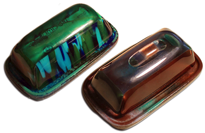 The two parts of a butter dish made from glass on a grey background. The base has a stick of butter on it. The dish is made from clear glass with white streaks in it.