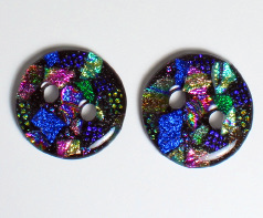 Two small round glass buttons on a gray background. The buttons each have two holes and are made of many smaller bits and pieces of dichroic-coated glass. The coating reflects in shades of yellow, green, blue, and purple.