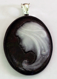 An oval glass cameo pendant on a light tan background. The body of the pendant is black. A womanâ€™s profile is filled in with opaque white on the pendantâ€™s right side, and the curve of her hair fits the curve of the pendant.