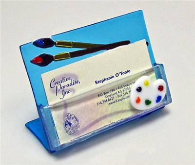 A business card holder made with light blue glass standing on a white background. The holder has two paintbrush decorations at the top and an artistâ€™s palette where the cards are held. The business cards inside are a cream color.