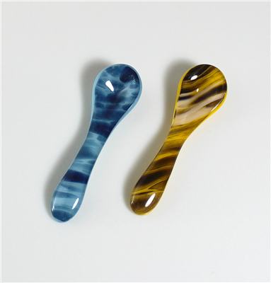 Two glass demitasse spoons on a grey background. The left spoon is dark blue with lighter blue patterning and the right spoon is a dark yellow with brown and cream striping.