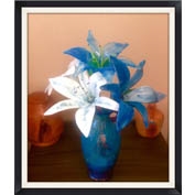 White and blue glass lilies in a blue vase. The lilies are realistic and include thin green stamens. The vase is on a brown table in front of a reddish wall, and the entire picture is surrounded by a black border.