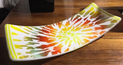 A glass rectangle curved at each end into a tray. The base glass is opaque white, and it has been decorated with a tie-dye style stencil radiating out from the center of the tray in shades of red, orange, and yellow. The tray is on a wooden table.