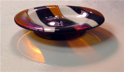 A glass dish with a round foot. The dish is made of alternating stripes of transparent amber, opaque cream, and opaque black glass. The amber has a slight iridescent shine. The dish is on a grey table with light shining through the transparent parts.