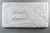 A long rectangular tile made of white ceramic. The carved texture on it reads Bon Appetit in script just to the left of center. The words are surrounded by bunches of grapes and leaves on vines.