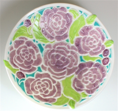 A top-down view of a circular glass dish. The dish has an opaque cream base with a peony design on it. The flowers are a light purple, the leaves a light green, and the spaces between them a vibrant turquoise.