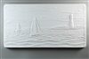 A long rectangular tile made of white ceramic. The carved texture shows a seascape with two sailboats on the waves. There is a peninsula to their right with a lighthouse, and the sky above them has a few long, wispy clouds.
