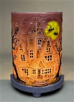 A glass rectangle bent into a semicircle and displayed on a purple base with a light behind it to create a lamp. The glass has a haunted house on it with a bright yellow moon and black trees. The glass is purple, but the light creates an orange gradient.