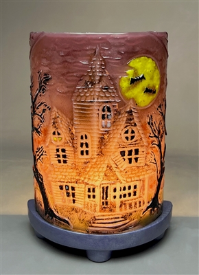 A glass rectangle bent into a semicircle and displayed on a purple base with a light behind it to create a lamp. The glass has a haunted house on it with a bright yellow moon and black trees. The glass is purple, but the light creates an orange gradient.