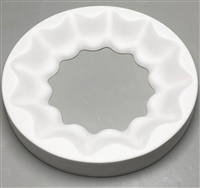 A circular white ceramic mold for fusing glass on a grey background. A scalloped circle has been cut out of the center. A larger scalloped circle surrounds it, and the edges of the larger circle slope downwards to meet the edges of the empty center.