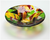 A small, circular bowl made from fused glass. The glass is made from many different colors and pieces all fused together into the bowl shape. It is primarily clear, green, red, orange, and yellow, with a few bits of black.
