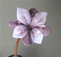 A fused glass flower displayed on a copper tube stem in front of a grey background. The flower has been shaped into an organic cup-like shape and has eight petals. Four are light purple and four are dark purple, and they alternate in placement.