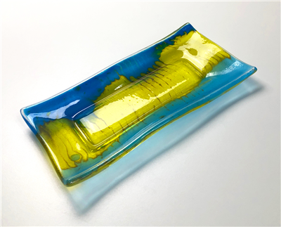 A rectangular dish made from fused glass. The edges are slightly waved and the center is a flat rectangle. Each long edge has been filled in a different shade of blue that has then been dragged into the yellow middle.