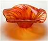 A side view of a bowl made from vibrant orange fused glass. The outer edges of the bowl are wavy, and the glass has a texture of many long lines spiraling in towards the deep center. Light shines through it, casting an orange shadow.