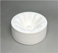A tall cylindrical white ceramic mold for fusing glass on a grey background. There is smaller fluted flower shape carved into it with a small circle at its base. It slopes inward like a bowl, and the flutes of the flower resemble petals.