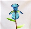 A fused glass iris flower on a copper tube stem. The iris is transparent blue with darker blue detail lines on the petals. There are small green leaves supporting the flower. And a couple more green leaves wrapped around the stem with wire.