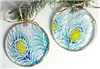 Two circular ornaments of fused glass. The base glass is clear, and they have each been decorated with the design of a peacock feather done in light blue, dark blue, and yellow. The edges of each ornament have been finished with liquid gold.