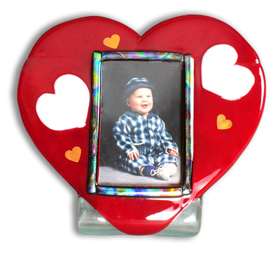 A fused glass picture frame made of a red heart attached to a bent clear bottom allowing it to stand. A rectangle of dichroic glass in the center of the heart frames a picture of a baby in plaid pajamas. Small white and yellow hearts are around the photo.