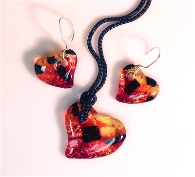 Three heart-shaped pendants made from fused glass. Each is made from pieces of red, orange, and yellow glass with a few chunks of black. The middle pendant is larger and strung from black cord. The two on the sides are smaller and on earring hangers.